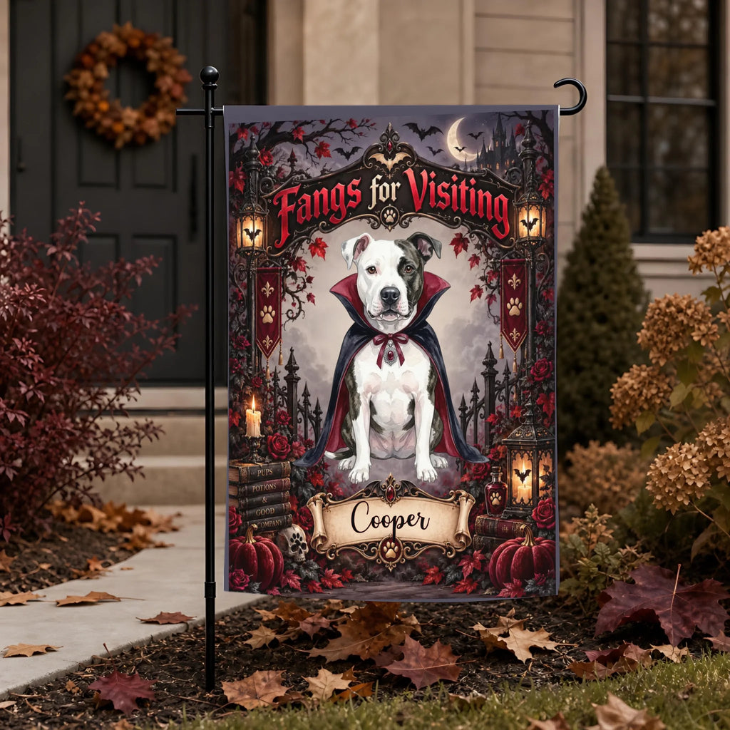 A Halloween-themed garden flag featuring a white pitbull-type dog dressed as a vampire with the text 'Fangs for Visiting' and the name 'Cooper' at the bottom, displayed on a black metal stand in a front yard with autumn leaves and landscaping.