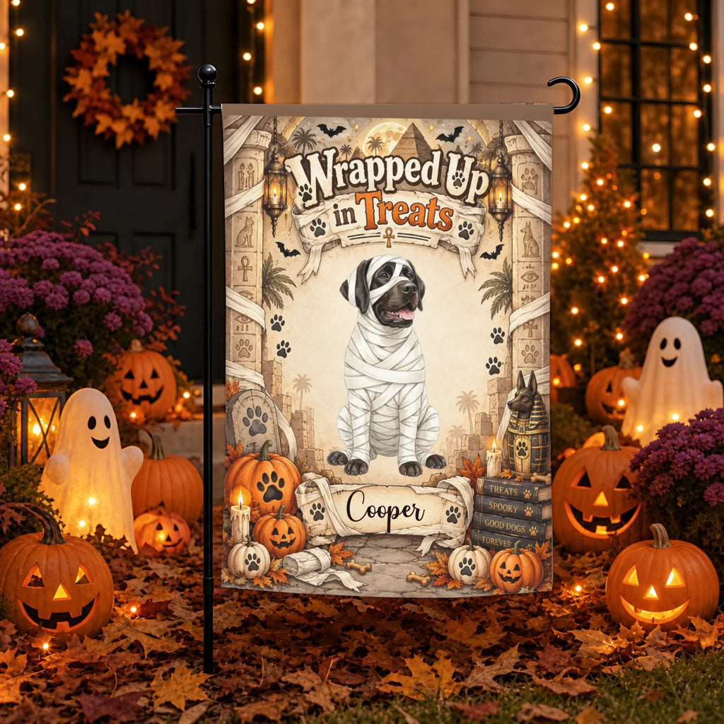 A Halloween-themed garden flag displayed on a black metal stand in a decorated front yard. The flag features a dog dressed as a mummy with the text 'Wrapped Up in Treats' and the name 'Cooper' at the bottom.