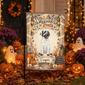 A Halloween-themed garden flag displayed on a black metal stand in a decorated front yard. The flag features a dog dressed as a mummy with the text 'Wrapped Up in Treats' and the name 'Cooper' at the bottom.