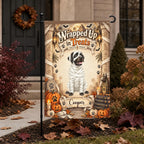 A decorative garden flag displayed on a black metal stand in a yard. The flag features a dog dressed as a mummy with the text 'Wrapped Up in Treats' and the name 'Cooper' at the bottom.