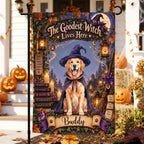 A decorative garden flag displayed on a black metal stand in a front yard. The flag features a golden retriever dressed as a witch with a purple hat and cape, surrounded by Halloween-themed elements including pumpkins, candles, a broomstick, and the text 'The Goodest Witch Lives Here' and 'Buddy'.