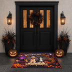 A Halloween-themed doormat featuring a witch and cat in a pumpkin patch, placed in front of a black front door decorated with a wreath, wall lanterns, pumpkins, and potted plants.