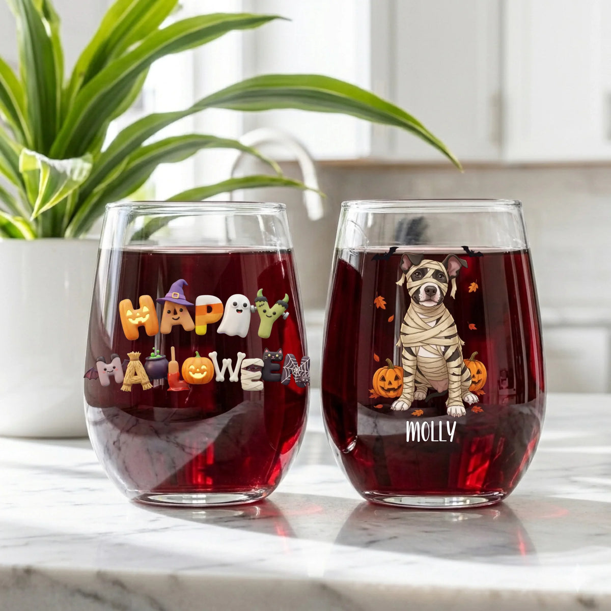 Two personalized Halloween wine glasses filled with red wine on a kitchen counter with a potted plant in the background