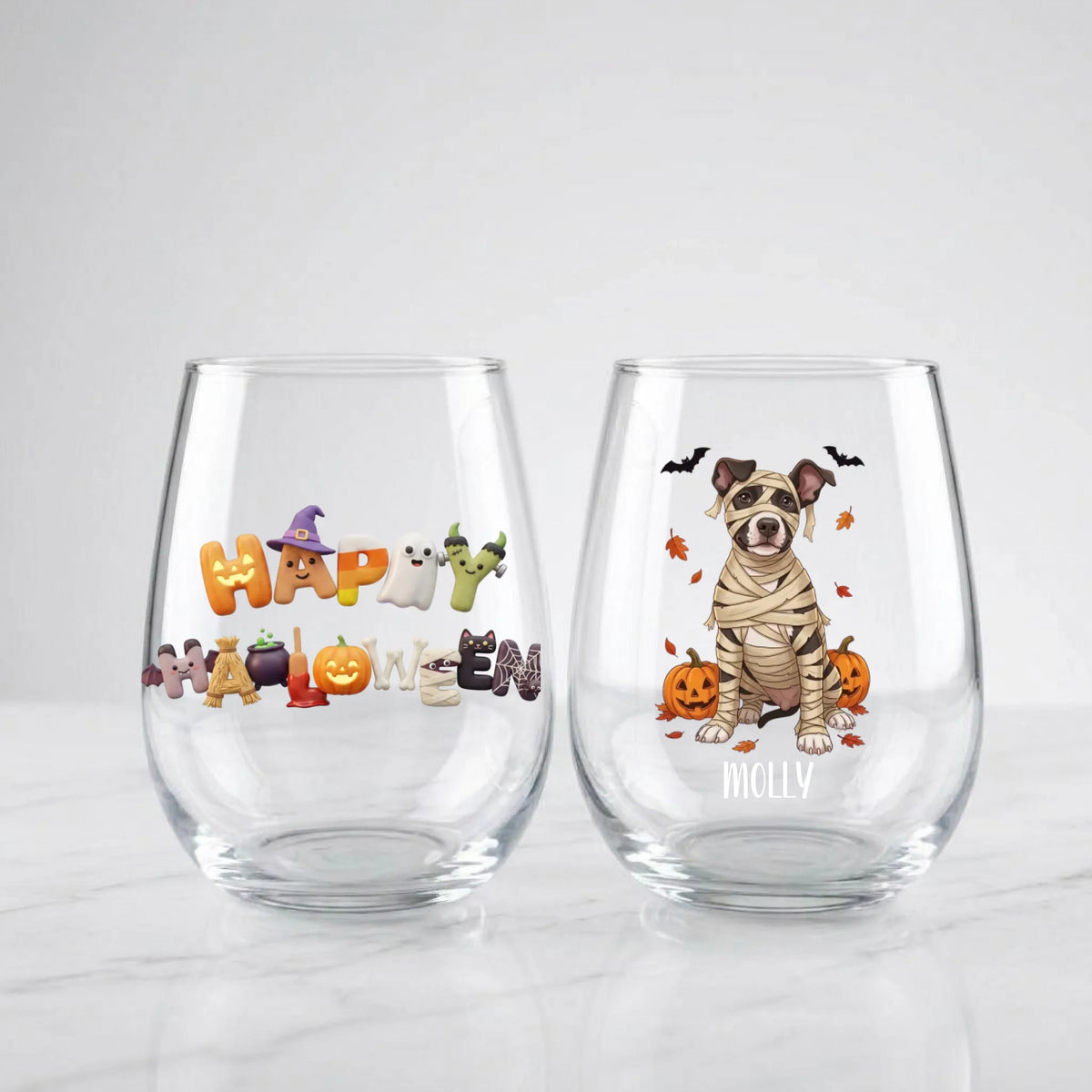 Two personalized Halloween stemless wine glasses on a marble countertop, one with 'Happy Halloween' lettering and one with a custom mummy-costume dog design and pet name