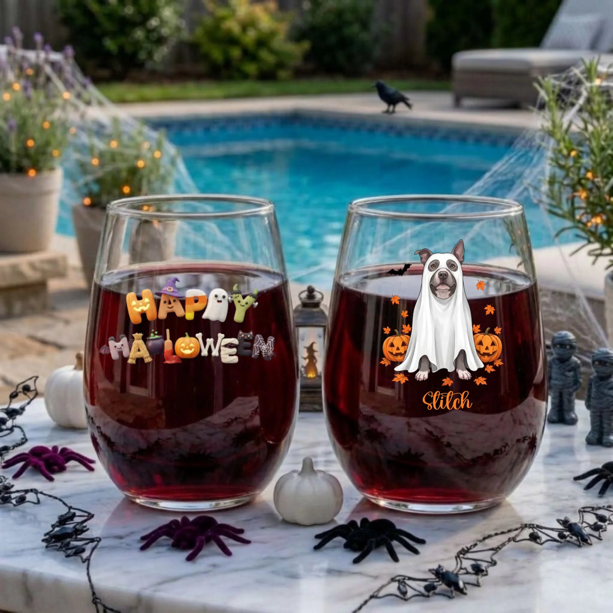 Two personalized Halloween stemless wine glasses filled with red wine on a poolside table, one with a ghost-costume dog with pumpkins