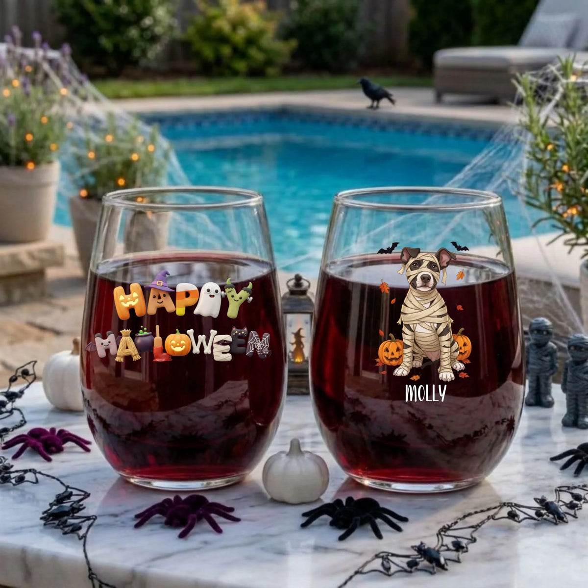 Two personalized Halloween stemless wine glasses filled with red wine on a poolside table, one with a mummy-costume dog with pumpkins