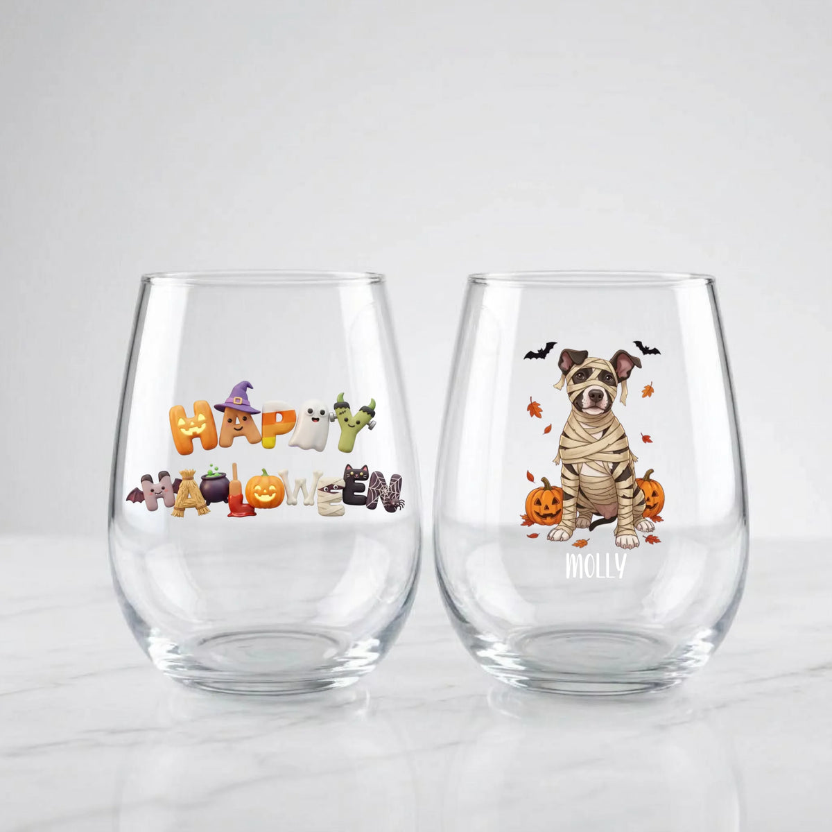 Two personalized Halloween stemless wine glasses on marble, one with 'Happy Halloween' lettering, one with a mummy-costume dog with pumpkins and pet name