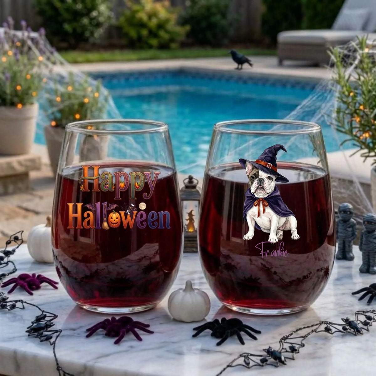 Two personalized Halloween stemless wine glasses filled with red wine on a poolside table, one with a watercolor witch-costume dog