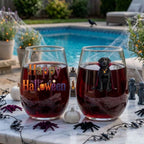 Two personalized Halloween stemless wine glasses filled with red wine on a poolside table, one with a watercolor vampire-costume dog