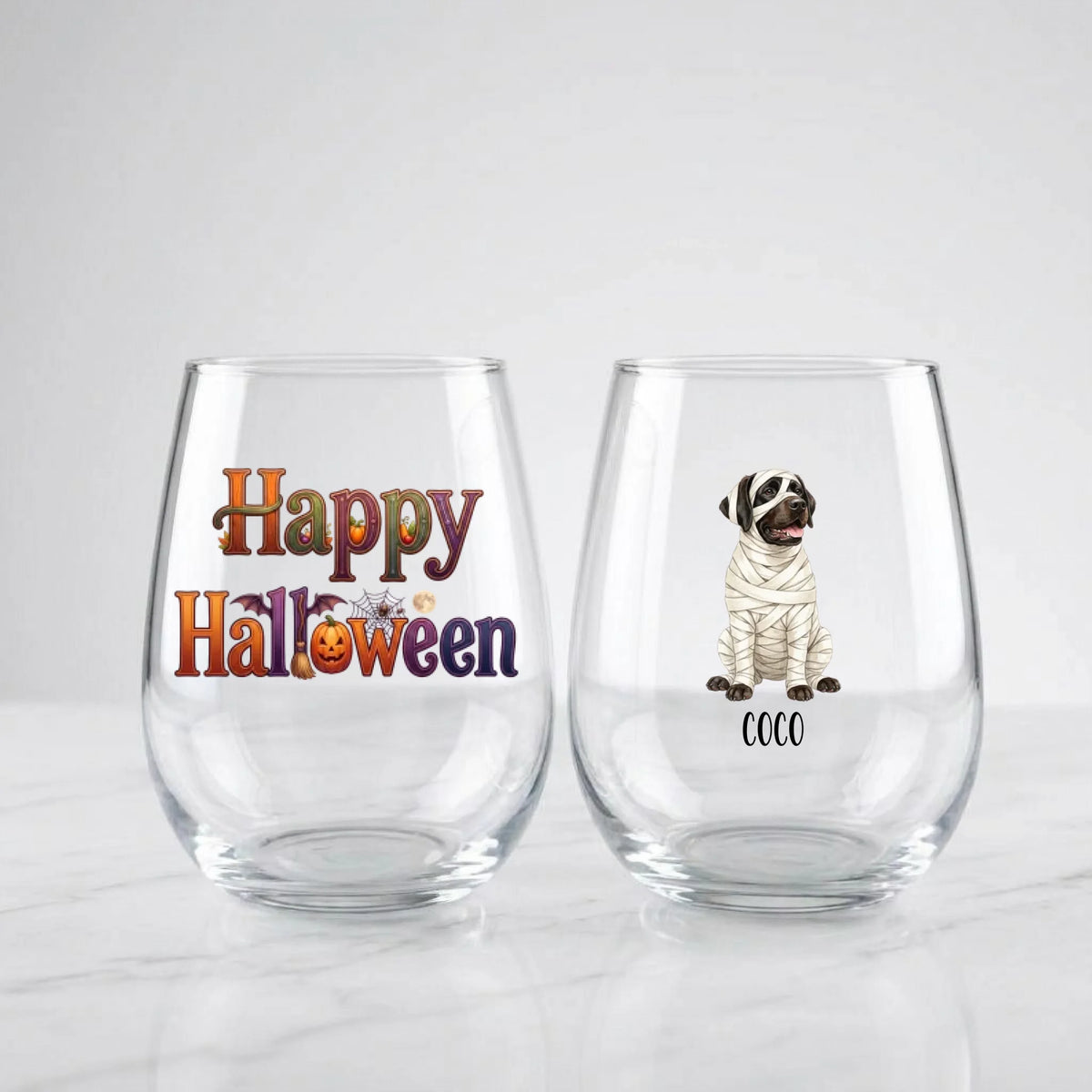 Two personalized Halloween stemless wine glasses on marble, one with 'Happy Halloween' lettering, one with a watercolor mummy-costume dog and pet name