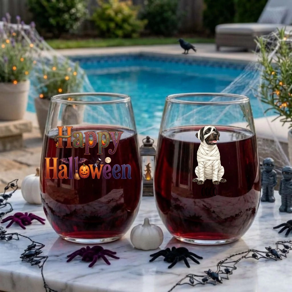Two personalized Halloween stemless wine glasses filled with red wine on a poolside table, one with a watercolor mummy-costume dog