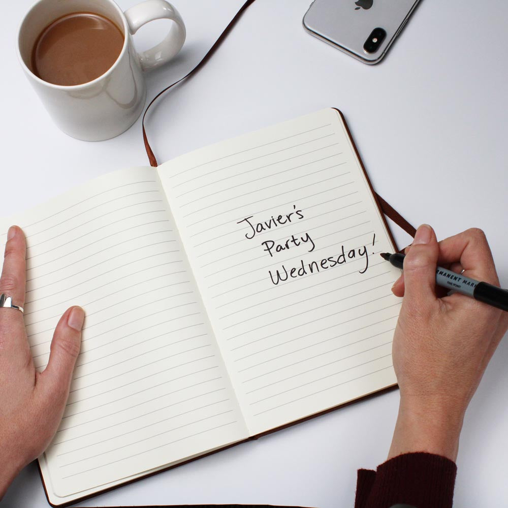 Person writing 'Javier's Party Wednesday!' in a notebook with a cup of coffee and phone on the side.